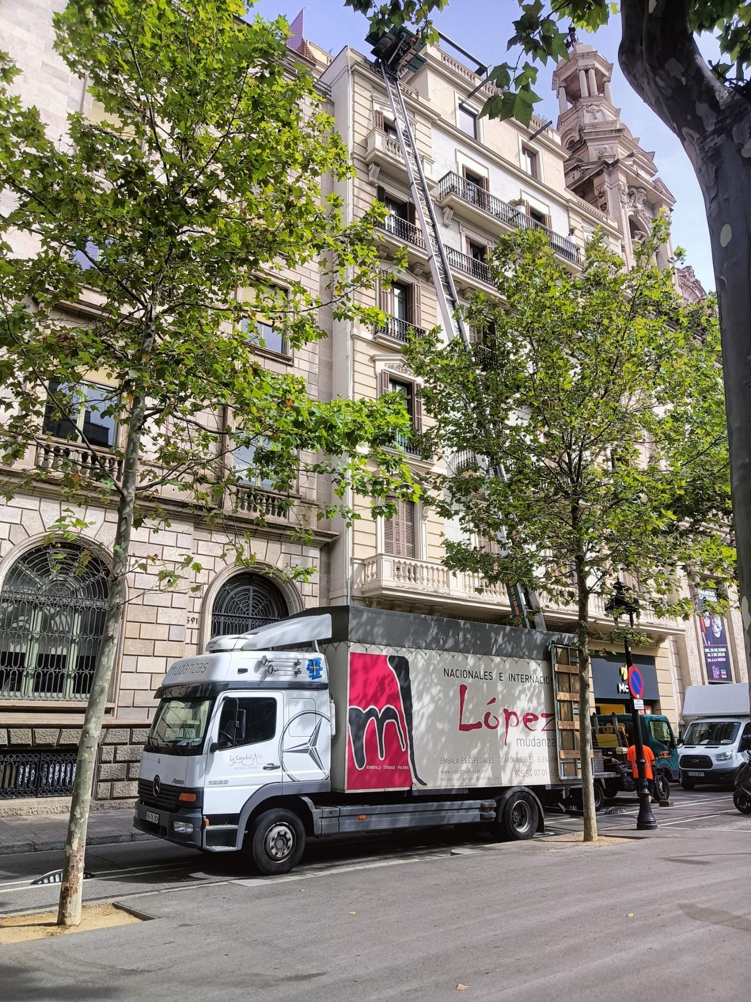 Camión con elevador al lado de un edificio de varios pisos, árboles en primer plano, personal de mudanzas presente.