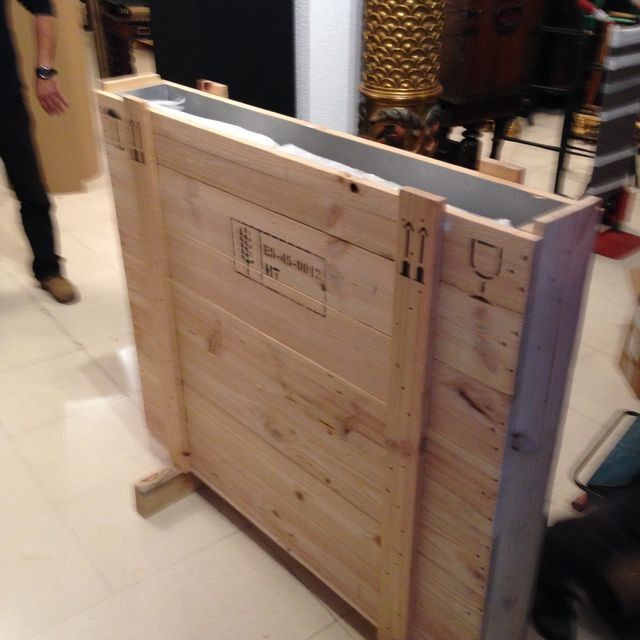Caja de madera, probablemente para envío, con símbolos y texto de envío. Persona parcialmente visible.