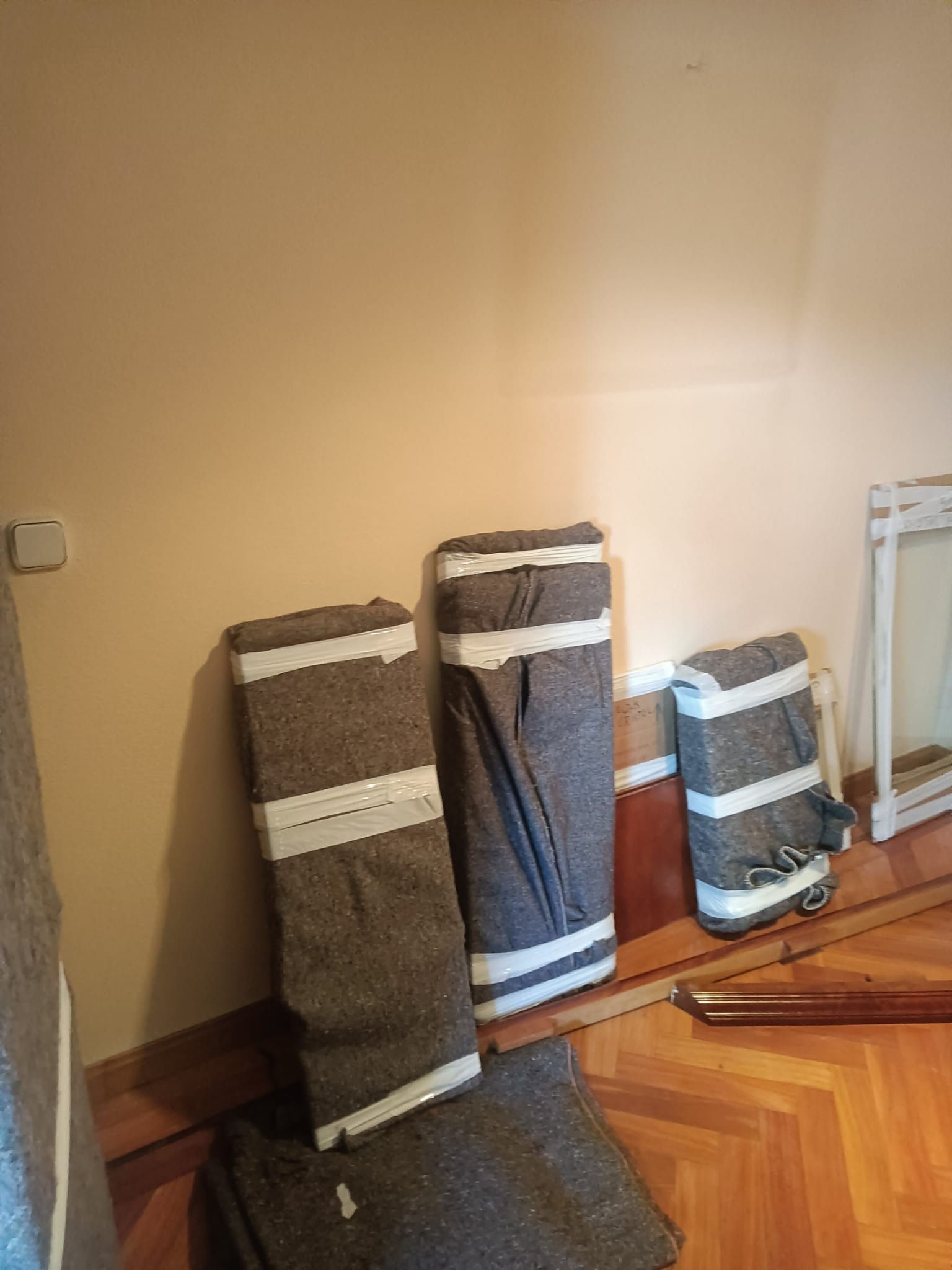 Alfombras enrolladas contra la pared, de distintos tamaños, sujetas con tiras blancas. Suelo de madera.