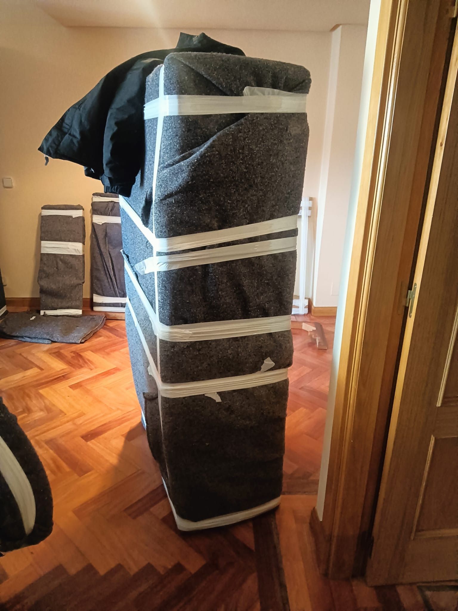 Un colchón gris envuelto y asegurado con cinta blanca se encuentra en posición vertical en una habitación con pisos de madera.