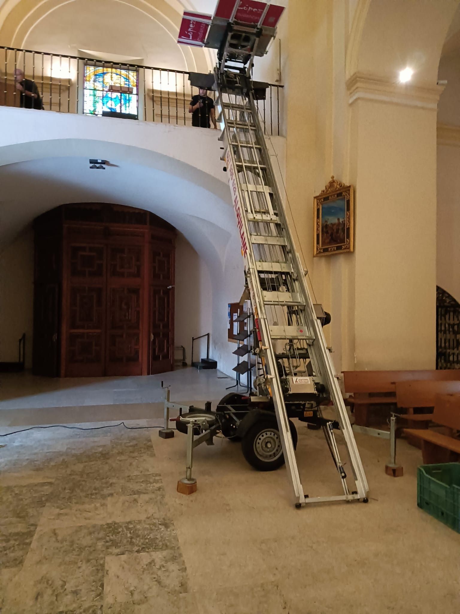 Un ascensor alto con ruedas dentro de una iglesia, utilizado para transportar objetos a un balcón; se ve una vidriera.