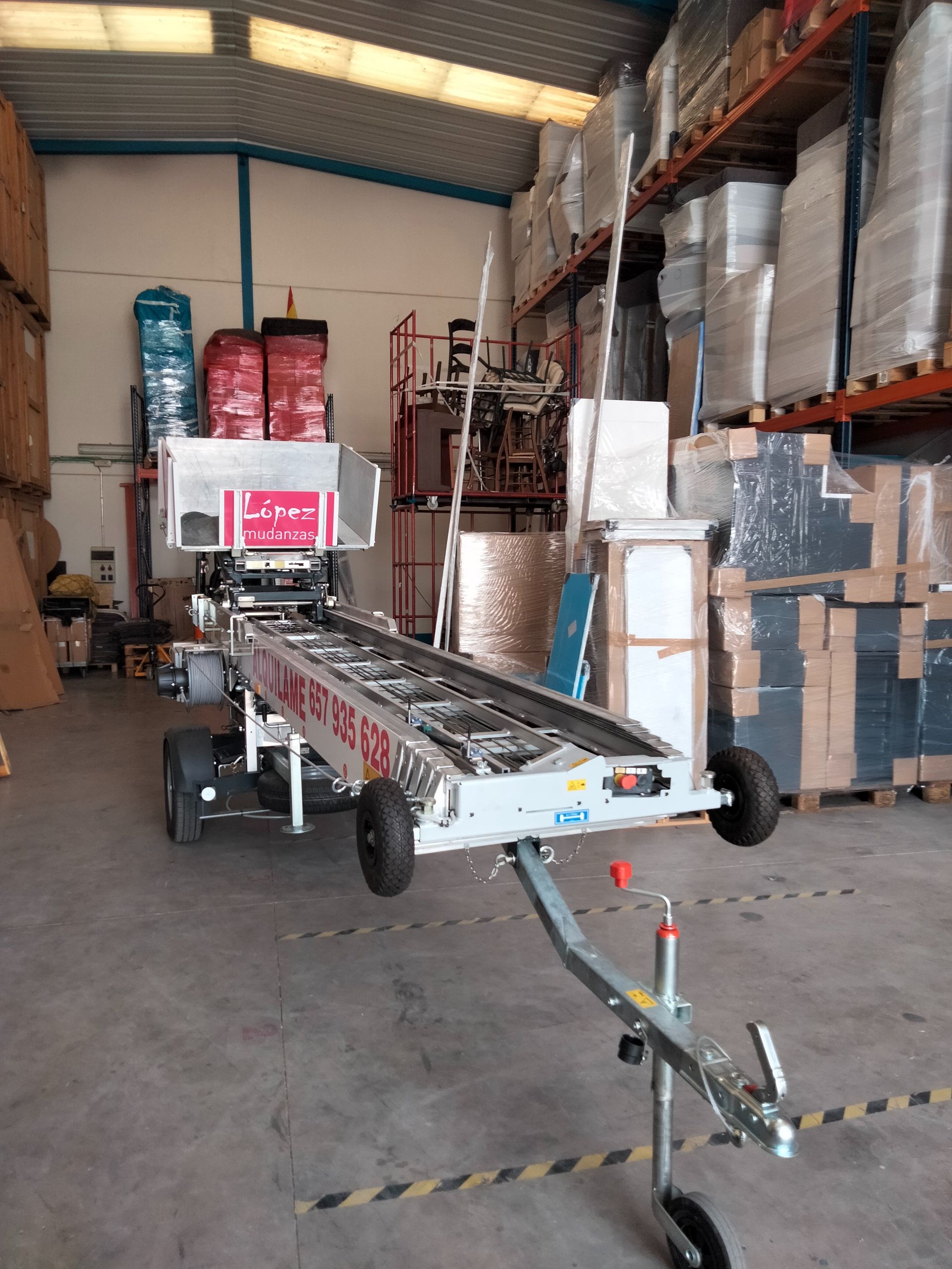 Almacén: Cinta transportadora plateada sobre un remolque. Neumáticos negros. Cajas y estanterías al fondo.