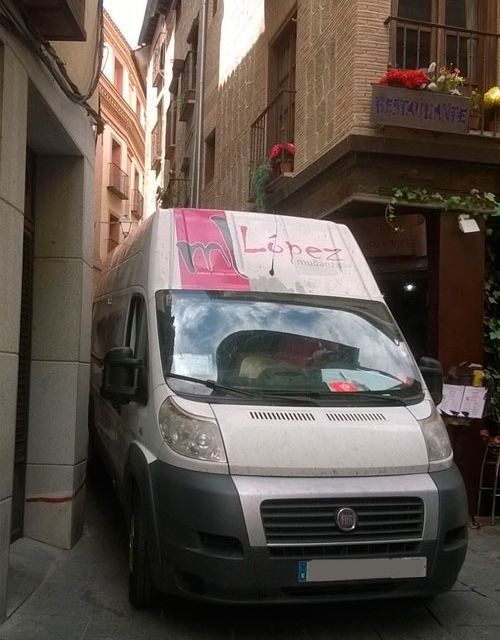 Furgoneta plateada con logo rosa, estacionada en un callejón estrecho, entre edificios.