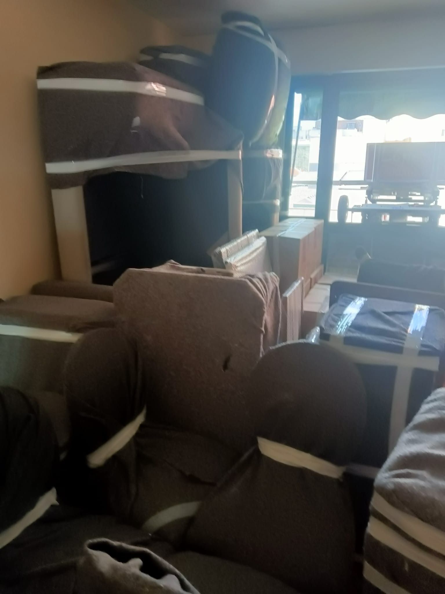 Muebles envueltos en fundas marrones y cinta adhesiva, cajas y una ventana visible, lo que indica mudanza.