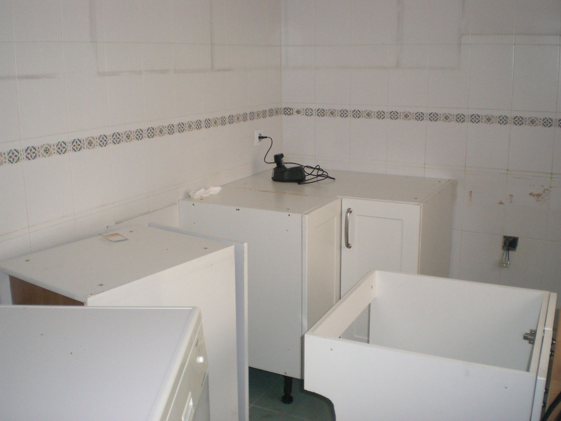 Gabinetes de cocina blancos en una esquina con gabinetes parcialmente instalados y protector contra salpicaduras de azulejos blancos.
