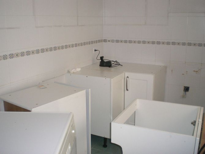 Gabinetes de cocina blancos en una esquina con gabinetes parcialmente instalados y protector contra salpicaduras de azulejos blancos.
