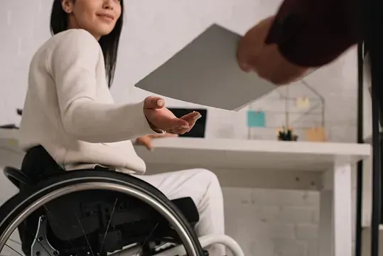 Mujer en silla de ruedas recibe documento de una persona en una oficina.