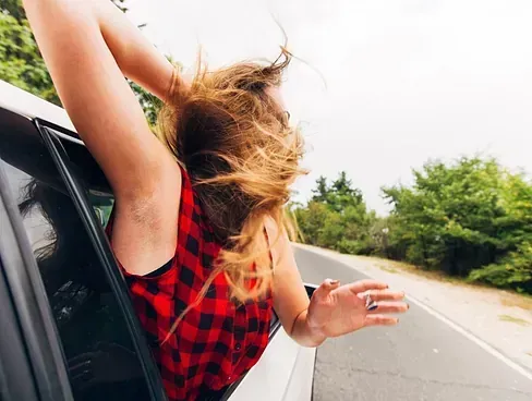 Una mujer con el cabello al viento se asoma por la ventanilla de un automóvil durante un viaje por carretera y sonríe con el brazo levantado.