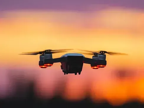 Dron volando al atardecer, con cielo naranja y morado. El dron tiene cuatro rotores con luces intermitentes.