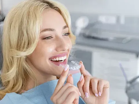 Una mujer rubia sonríe, sosteniendo un alineador transparente en sus dedos, en una silla dental.