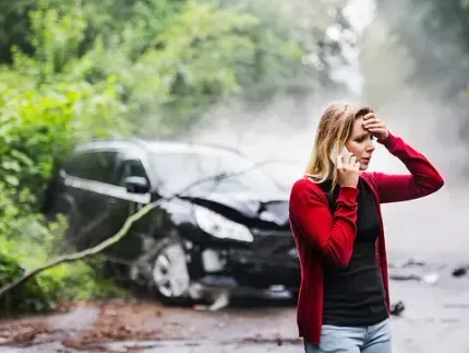 Una mujer con un cárdigan rojo habla por teléfono, con aspecto angustiado, está parada cerca de un automóvil negro dañado en una carretera mojada después de un accidente automovilístico.