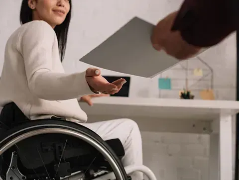 Mujer en silla de ruedas recibe papeles de una persona, sentada en un escritorio en un entorno de oficina.