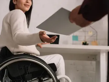 Mujer en silla de ruedas recibe papeles de una persona, sentada en un escritorio en un entorno de oficina.