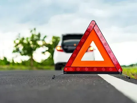 Triángulo de emergencia en una carretera, con un coche al fondo y el maletero abierto, indicando una avería.
