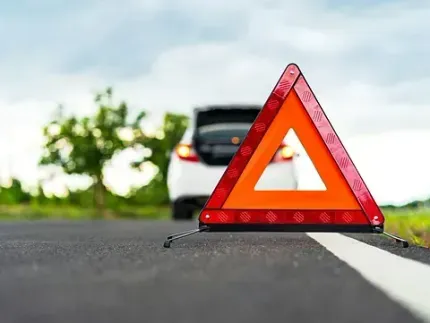 Triángulo de emergencia en una carretera, con un coche al fondo y el maletero abierto, indicando una avería.
