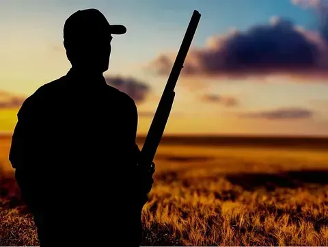 Silueta de una persona sosteniendo una escopeta, de pie en un campo al atardecer.