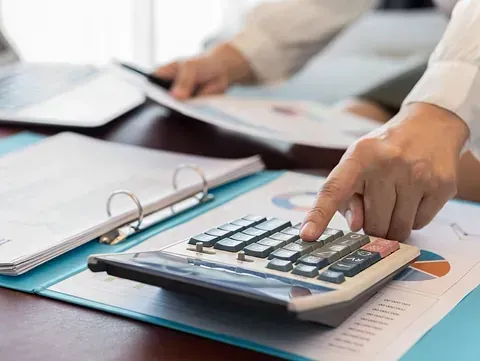 Las manos de una persona usando una calculadora en un escritorio con documentos financieros y una computadora portátil.