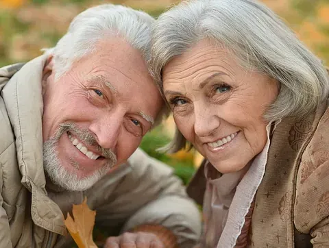 Pareja de ancianos sonriendo, con las cabezas juntas, en un entorno otoñal; el hombre tiene barba blanca.