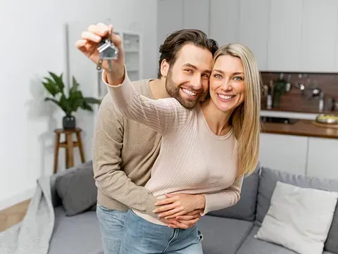 Una pareja se abraza en su nuevo hogar, sonriendo y sosteniendo las llaves. Están junto a un sofá gris en una sala de estar luminosa.
