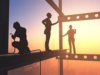 Obreros de la construcción recortados contra una puesta de sol, soldando e inspeccionando vigas de acero en el marco de un edificio de gran altura.