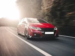 Un coche deportivo rojo circula a toda velocidad por una carretera con la luz del sol al fondo y árboles a los lados.