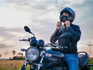 Hombre en motocicleta ajustándose el casco. Lleva chaqueta negra y vaqueros azules. Al fondo, el atardecer.