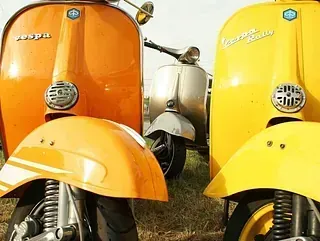 Dos scooters Vespa clásicas, de color naranja y amarillo, aparcadas una al lado de la otra, con una plateada al fondo.