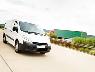 Furgoneta de reparto blanca circulando a toda velocidad por la autopista, con un camión remolque verde al fondo.