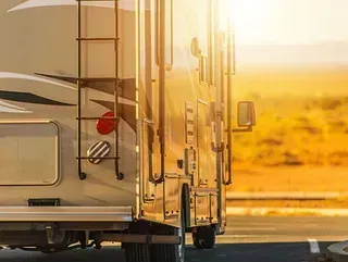 Parte trasera de una autocaravana estacionada al aire libre, bañada por la cálida luz del sol. Cuenta con escalera, compartimentos de almacenamiento y espejo lateral.