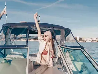 Una mujer en un barco, levantando el brazo en señal de celebración. Lleva gafas de sol y una bufanda, con el mar y una ciudad al fondo.