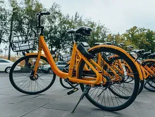 Bicicletas de alquiler de color naranja estacionadas en una acera en un entorno al aire libre, con un fondo arbolado y automóviles visibles.