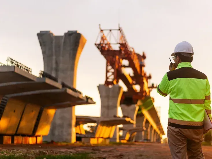 Trabajador de la construcción con chaleco de alta visibilidad y casco usando una radio, supervisando un puente en construcción al atardecer.
