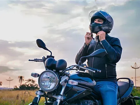 Un hombre en motocicleta se ajusta el casco, preparándose para conducir. Lleva una chaqueta negra y vaqueros azules contra un cielo nublado.