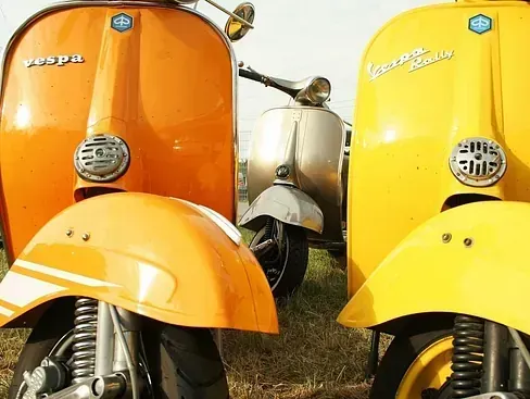 Primer plano de tres scooters Vespa, naranja, amarilla y plateada, estacionadas en el césped.