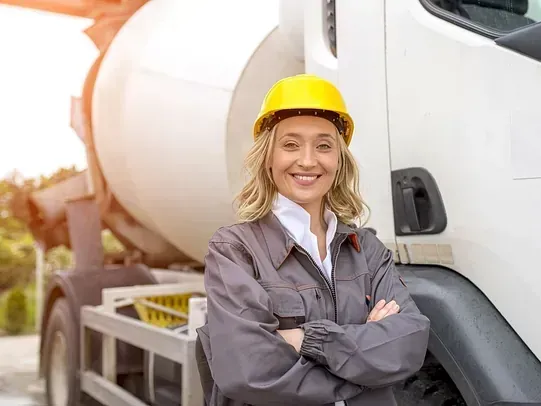 Una mujer con casco amarillo y traje de trabajo gris sonríe, con los brazos cruzados, parada frente a un camión hormigonera blanco.