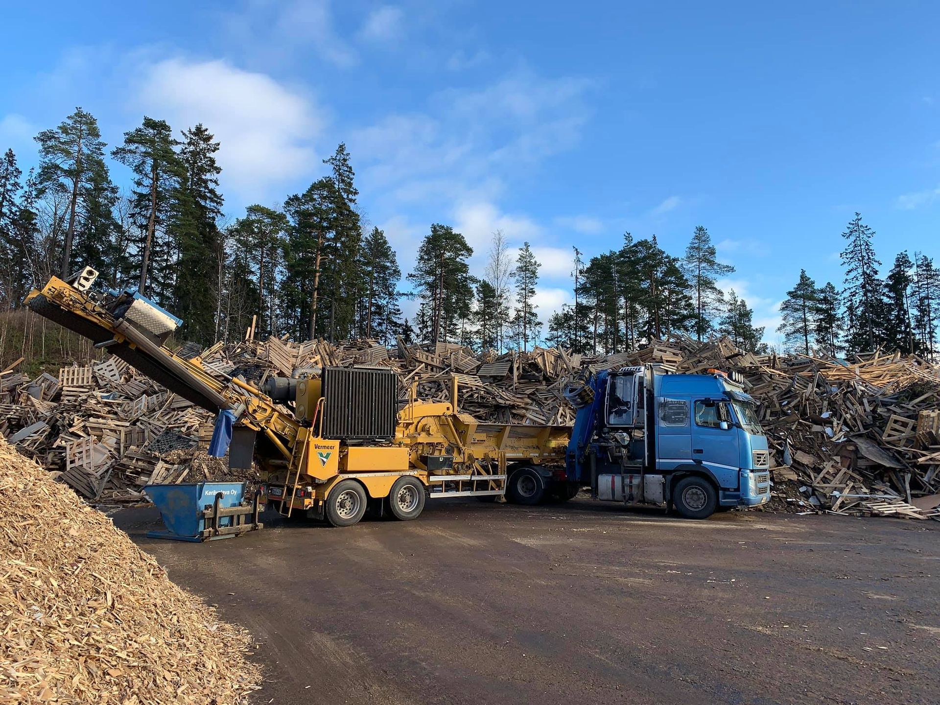 Sininen kuorma-auto ja keltainen työkone pysäköitynä hakekasan edessä.