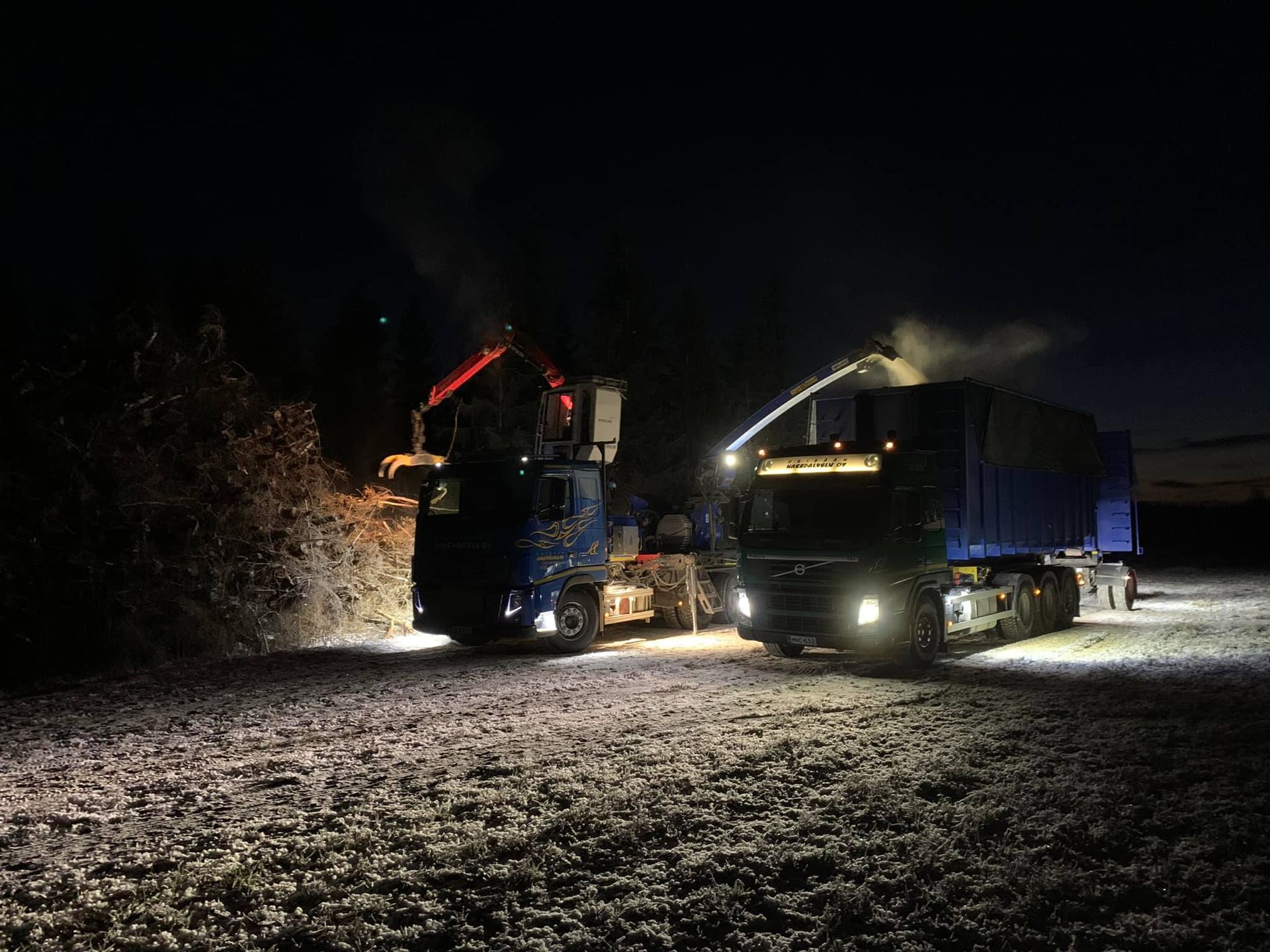 Kaksi kuorma-autoa nostimilla lumisella tiellä yöllä.