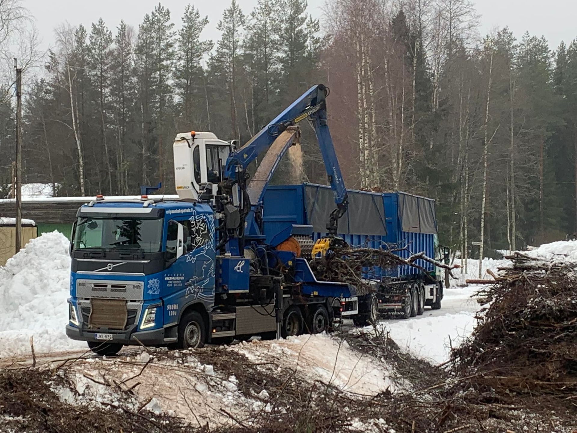 Sininen kuorma-auto nostimella, kyytiin nostetaan puun runkoja 