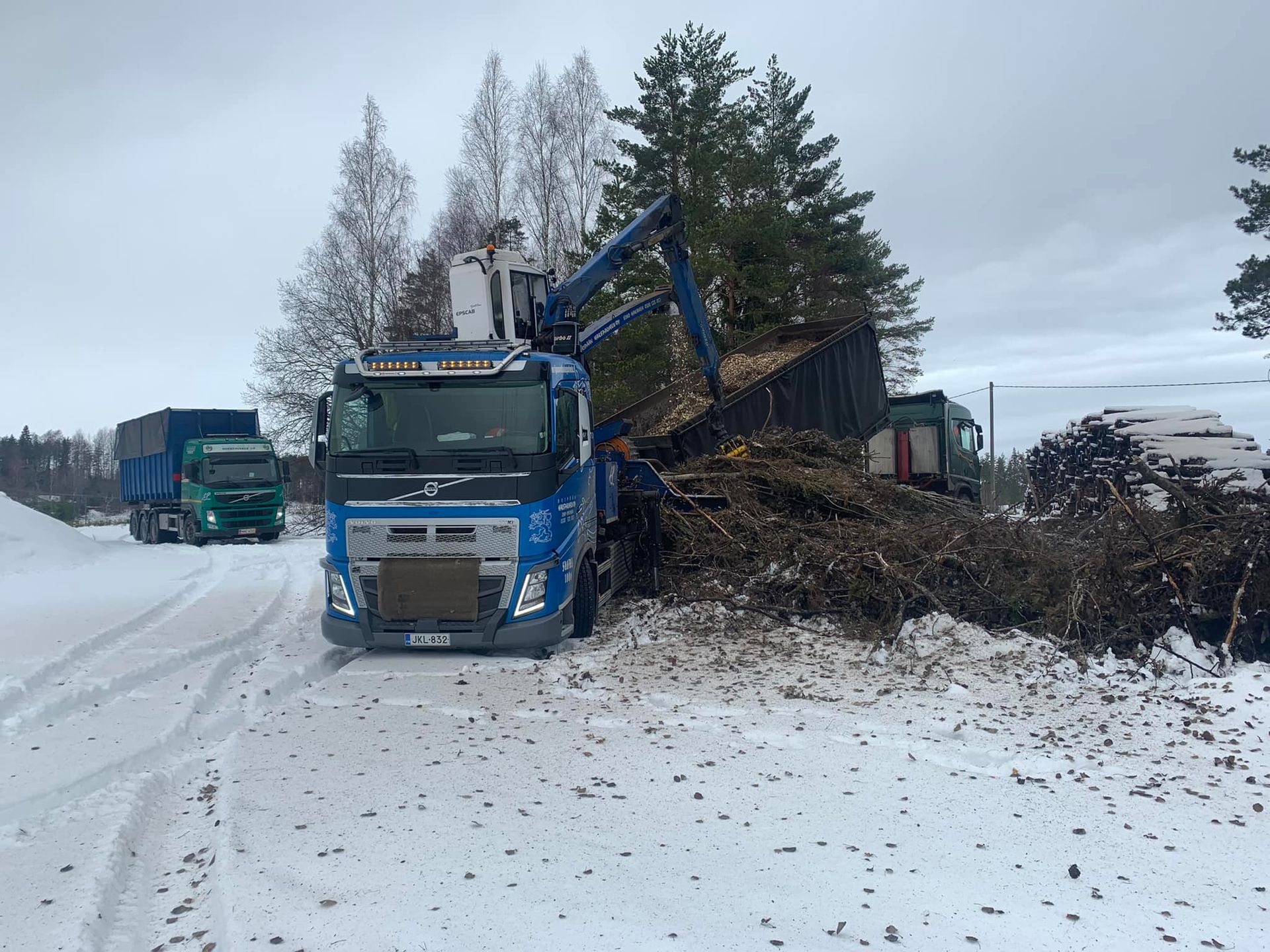 Sininen kuorma-auto lastattavana, vihreä kuorma-auto taustalla.