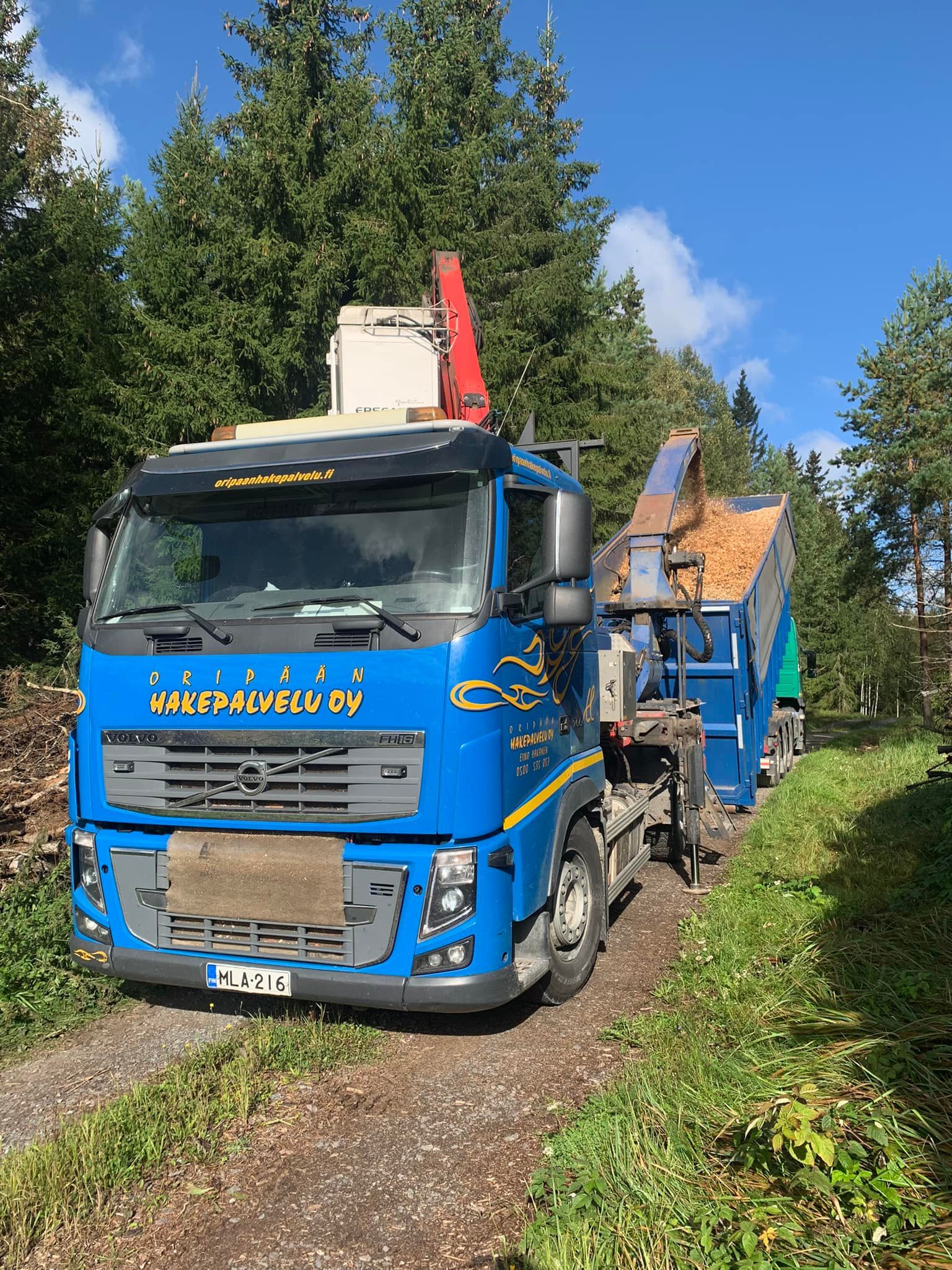 Sininen kuorma-auto on pysäköity hiekkatien viereen, lastina haketta