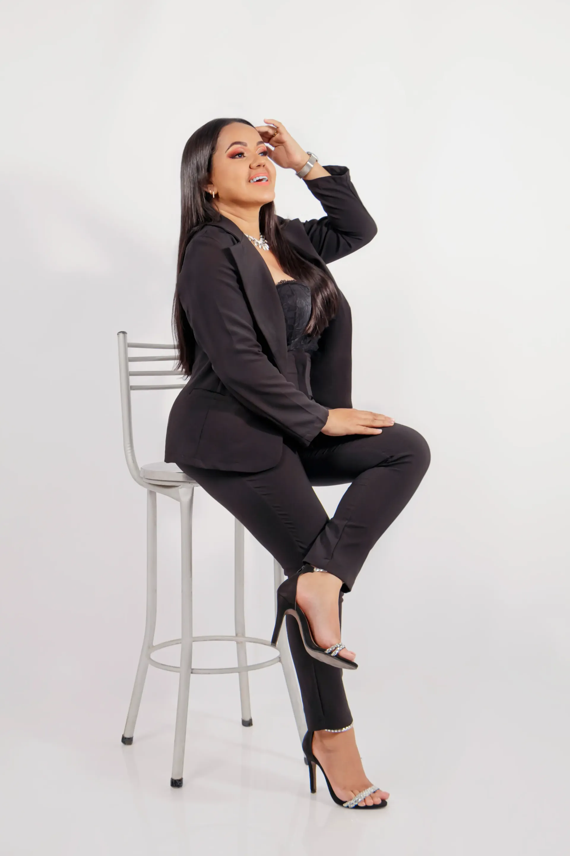 Mujer con chaqueta y pantalón negros sentada en un taburete de metal sobre un fondo blanco, posando con una mano en la frente.