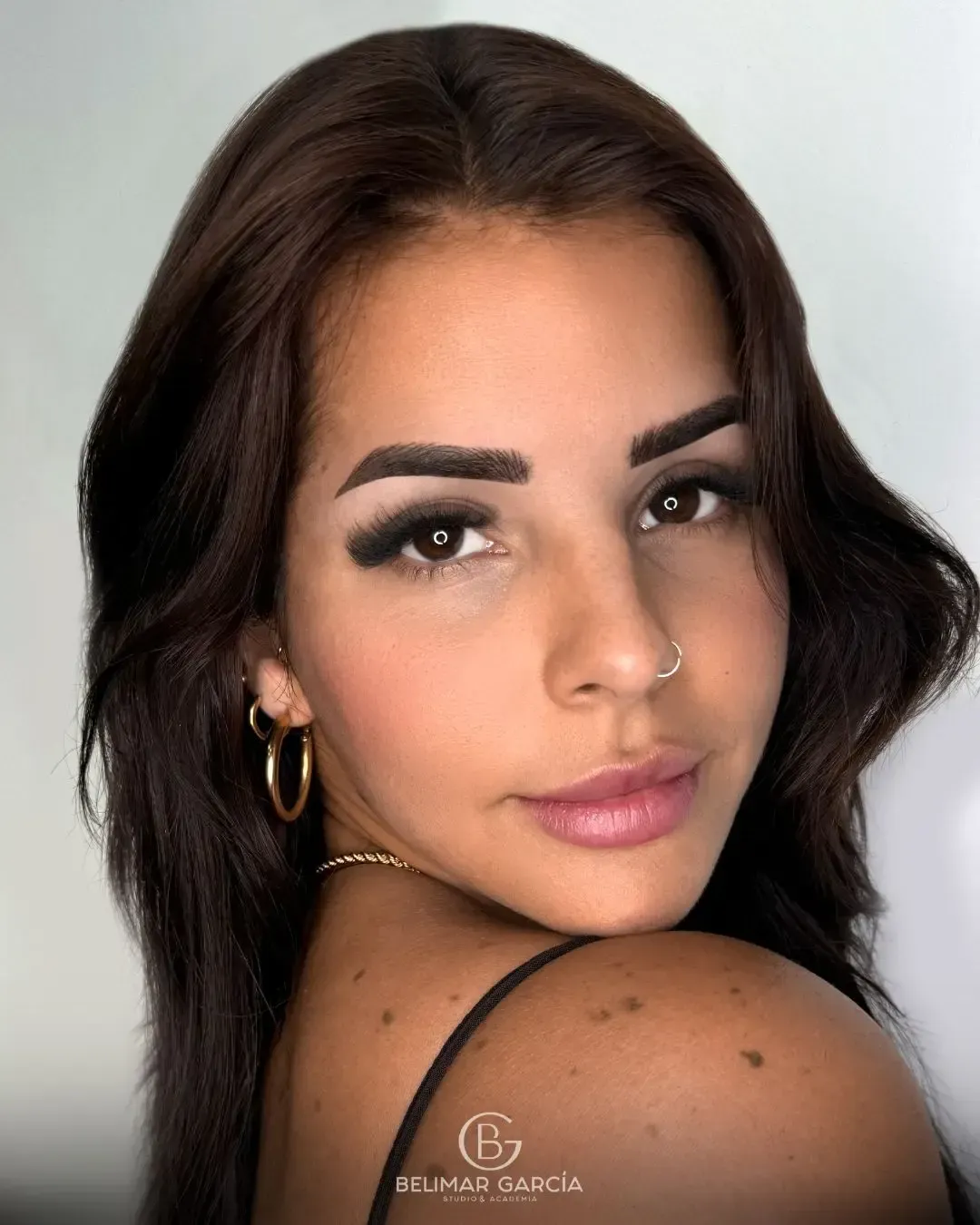 Retrato de cerca de una persona con cabello corto y oscuro, pendientes de aro dorados y un piercing en la nariz sobre un fondo claro.