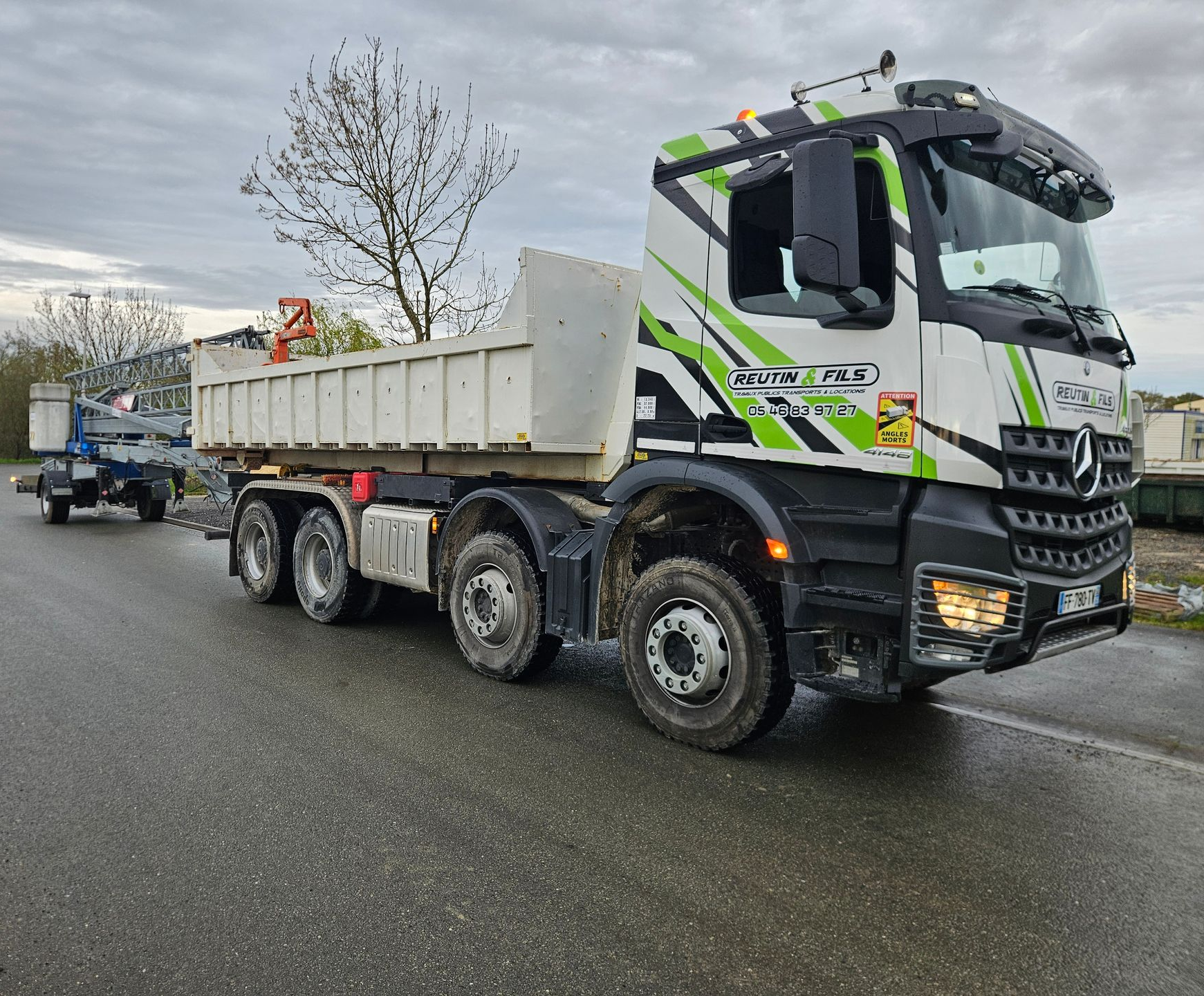 Un camion-benne blanc et vert sur une route mouillée, tirant une remorque à benne basculante.