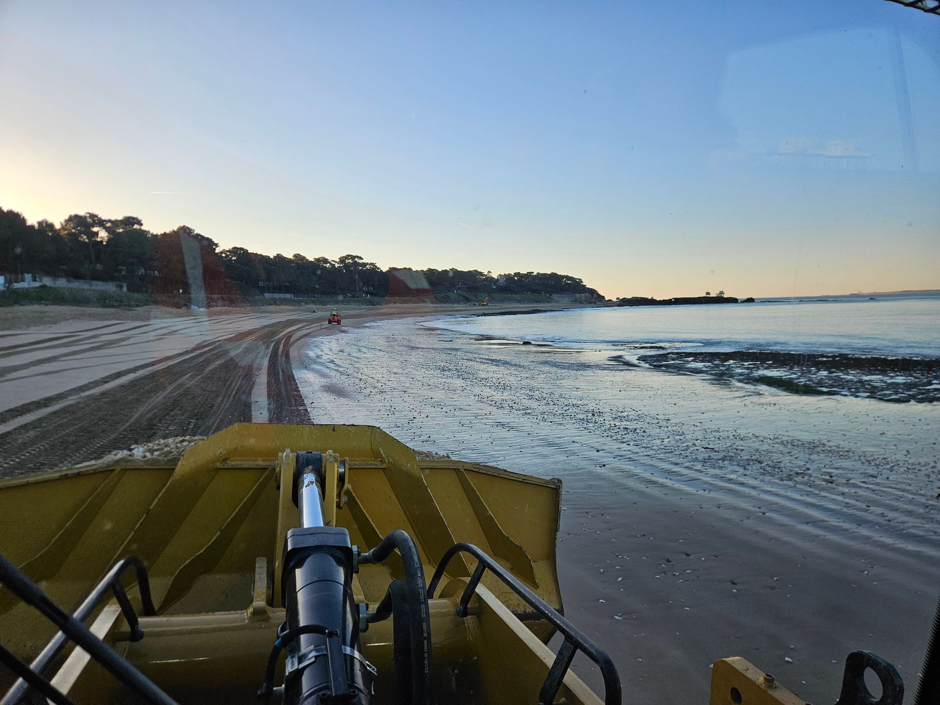 Un bulldozer jaune aplanit le sable d'une plage près de l'eau, avec un ciel de coucher de soleil en arrière-plan.