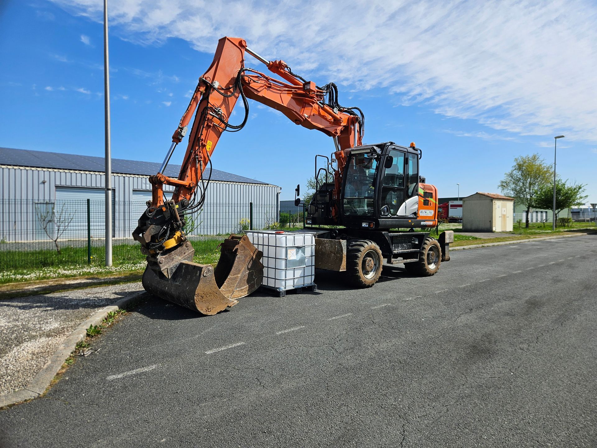 Une excavatrice orange équipée d'une pince de préhension tient une bûche et une palette sur une route asphaltée.