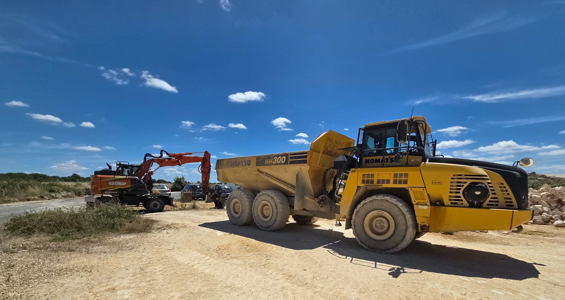 Un camion-benne jaune et une excavatrice sur un chantier de construction sous un ciel bleu nuageux.