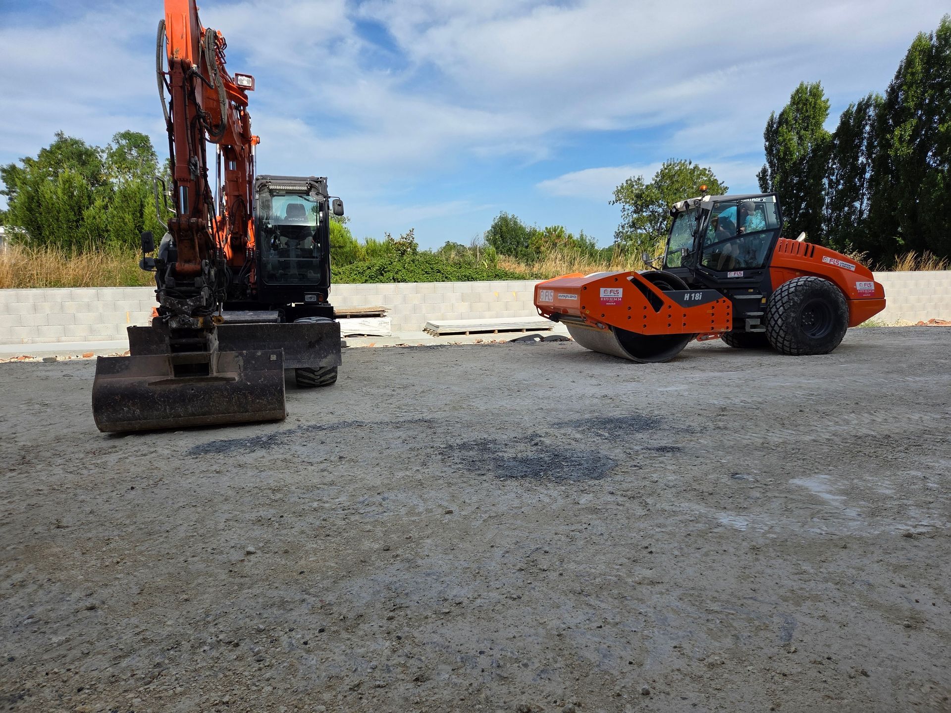Une excavatrice et un rouleau compresseur compactent une surface de gravier sur un chantier de construction.