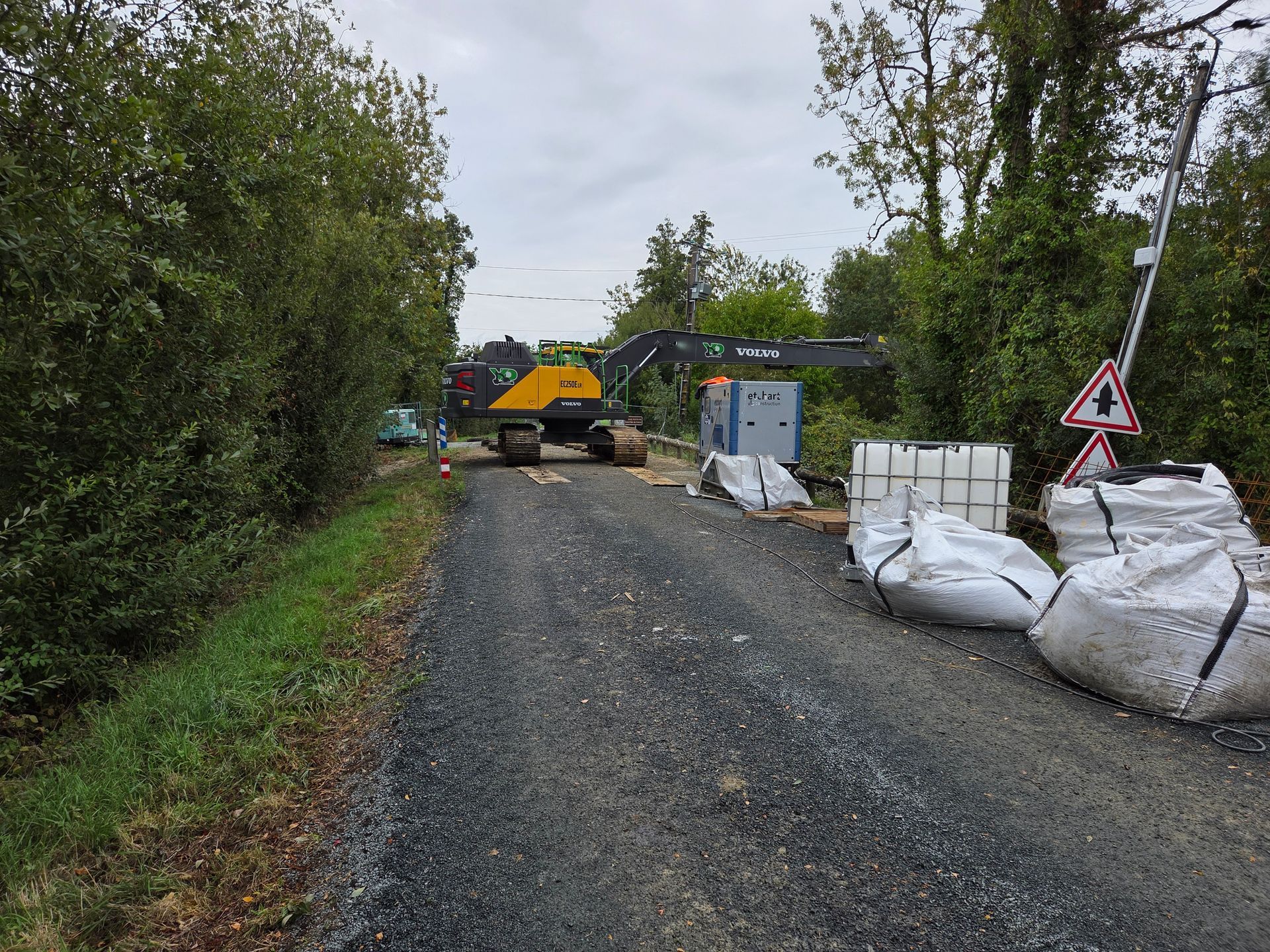 Route de gravier avec des engins de chantier : une excavatrice jaune, des conteneurs blancs et un panneau de signalisation.