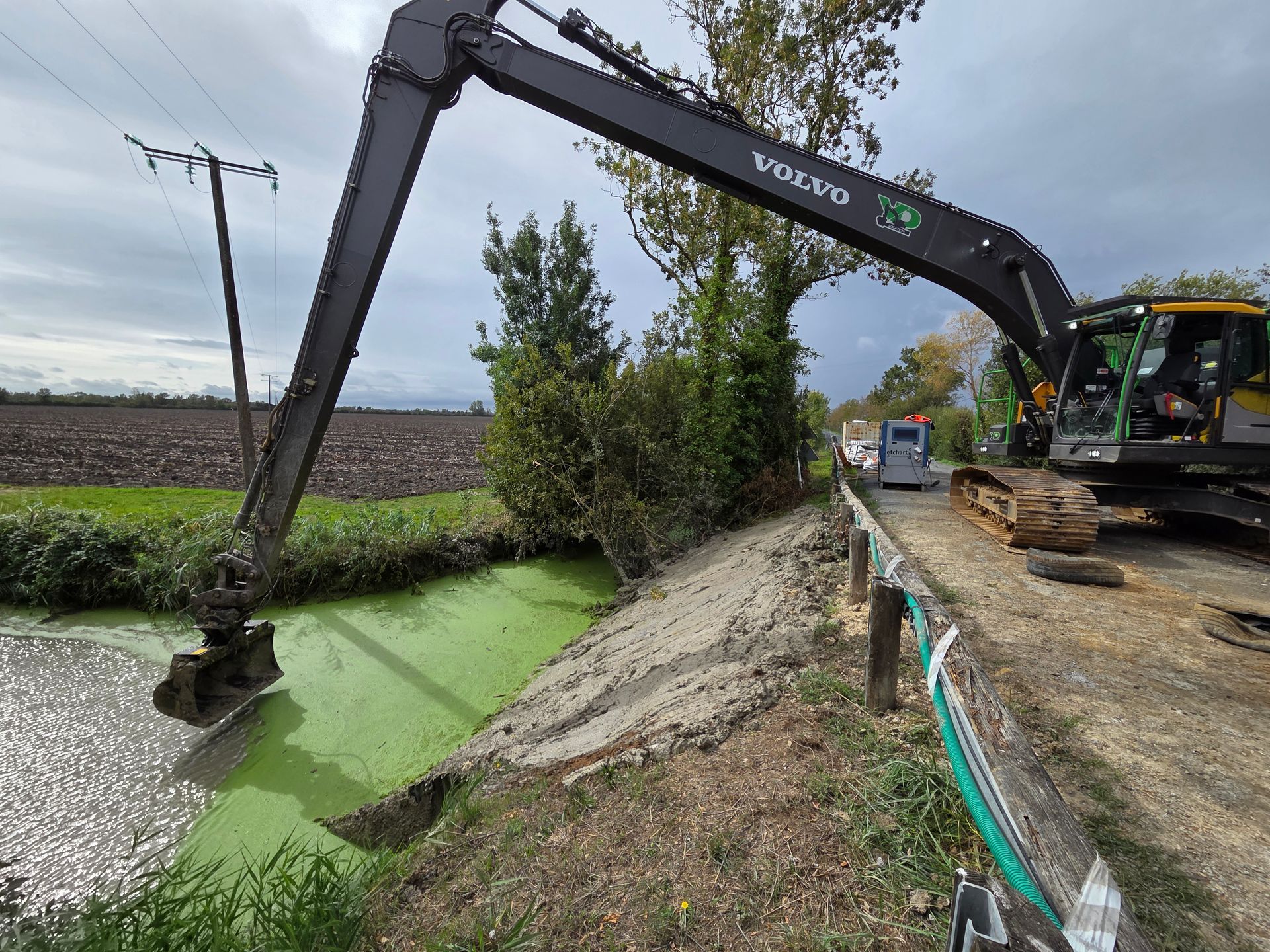 Une pelle mécanique Volvo dégage des débris d'un fossé rempli d'algues vertes, en bordure d'une route de campagne.