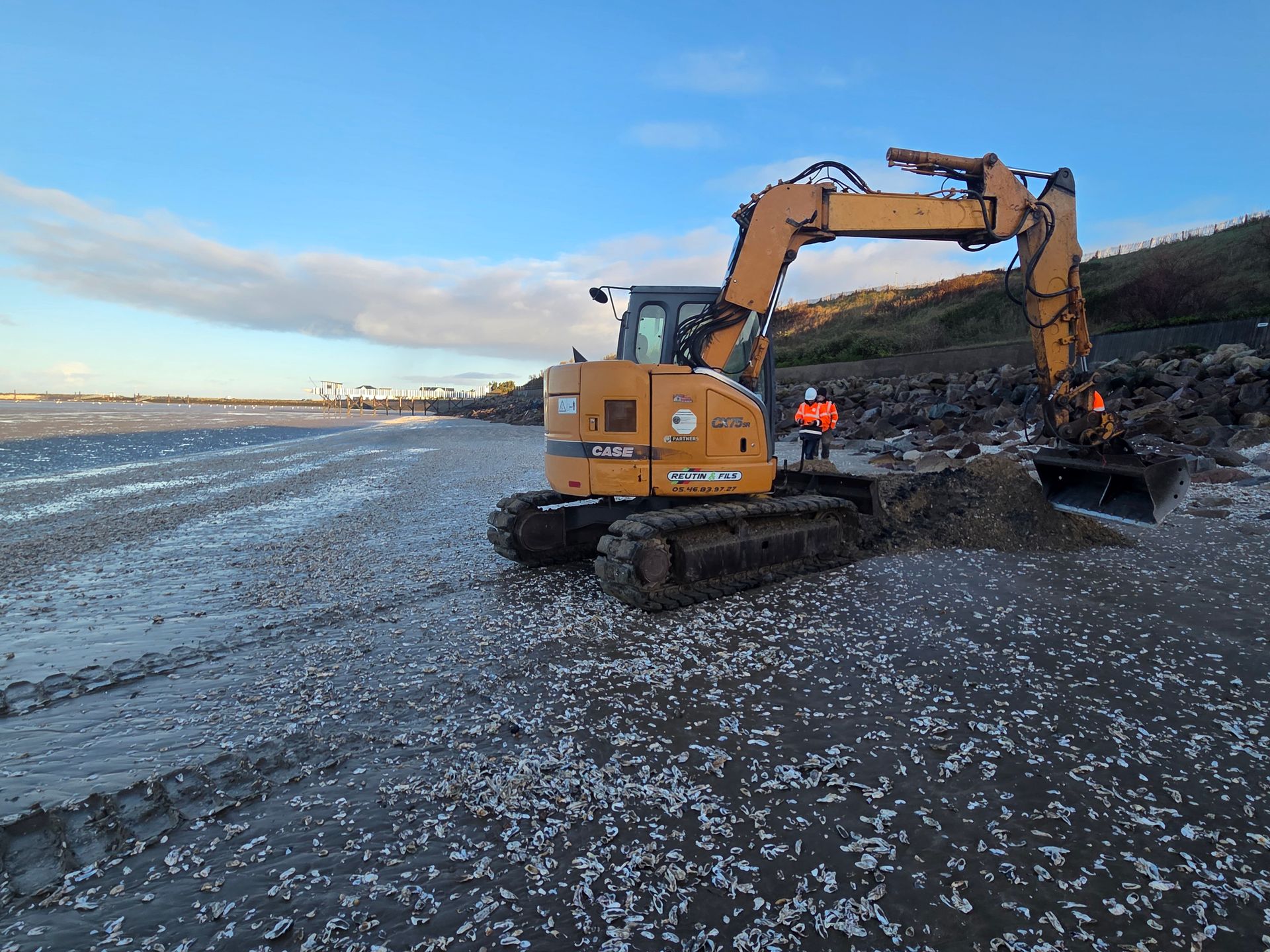 Une pelleteuse sur une plage, en train de ramasser des débris. Deux personnes en gilets orange se tiennent à proximité, sous un ciel bleu.